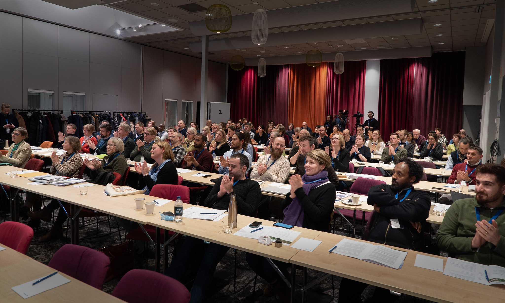 A big audience sitting at long tables clapping their hands.